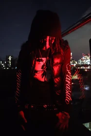 A black beanie and puffer vest under red light against the night skyline.