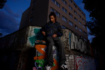 A seated look in a structured denim jacket beside a graffiti-covered wall.